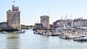 Um porto com barcos atracados, um castelo com uma bandeira e edifícios ao fundo.