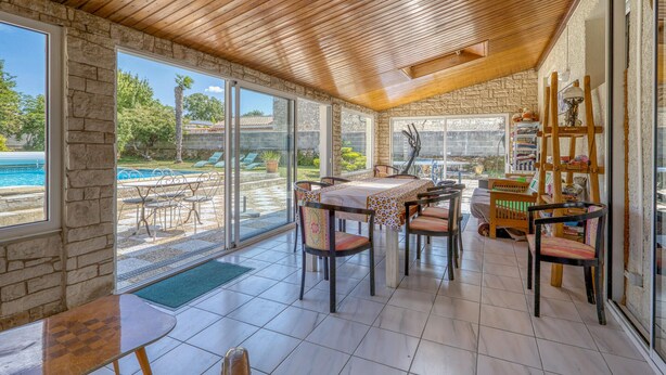 Um quarto banhado pelo sol, com teto de madeira, janelas amplas, piscina lá fora e uma área de jantar com cadeiras e mesa.