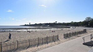 Uma praia com cerca de madeira, calçada de concreto e vista distante de edifícios e árvores.