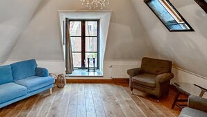 A loft-style room with a sloped ceiling, a large window with a view of buildings, a blue sofa, a brown armchair, and a small table with a candle.