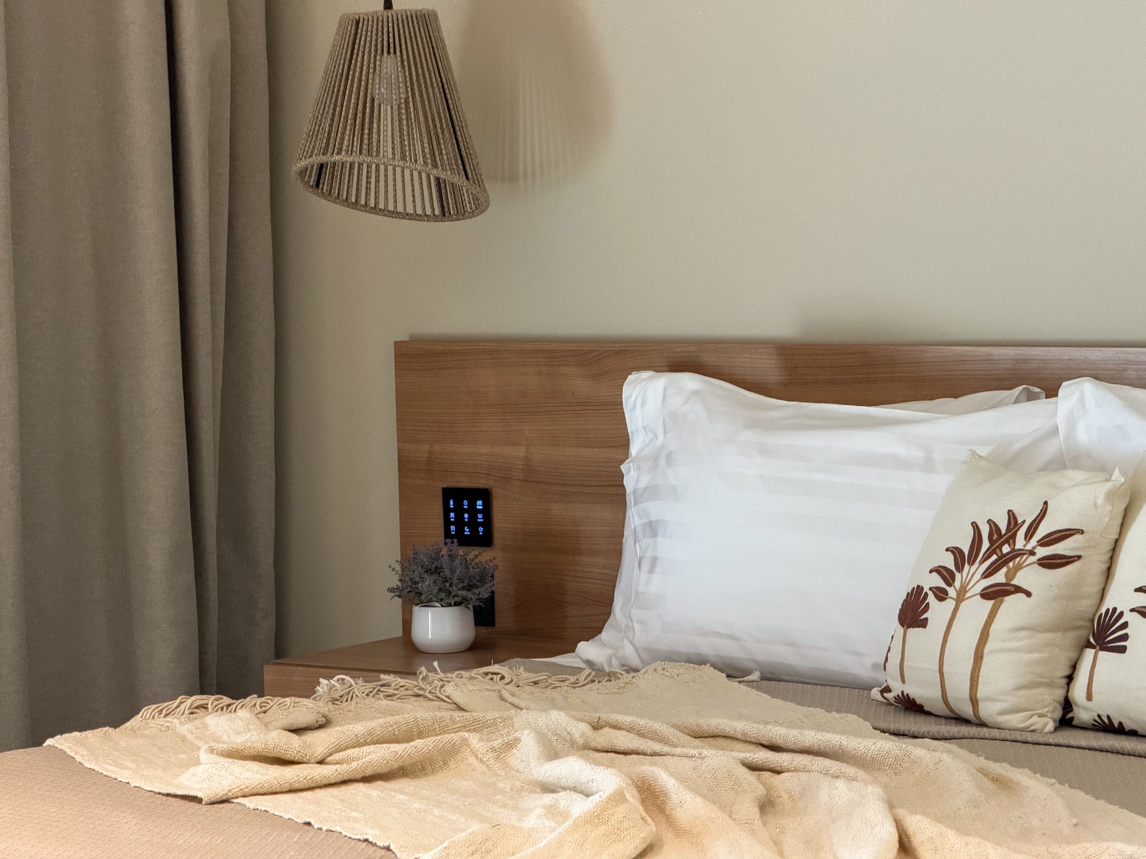 A neatly made bed with a white pillow and a decorative pillow with a floral pattern, a bedside table with a small potted plant, and a wall-mounted control panel.