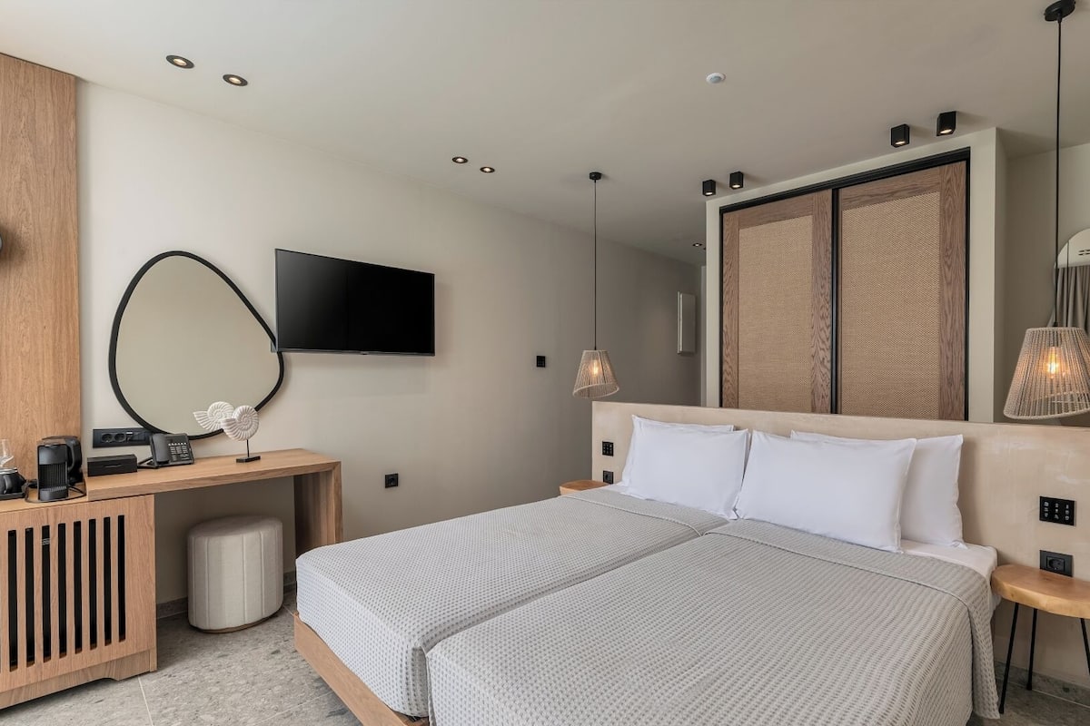 A modern hotel room with a bed, bedside tables, a desk with a computer, a TV, and a large mirror.