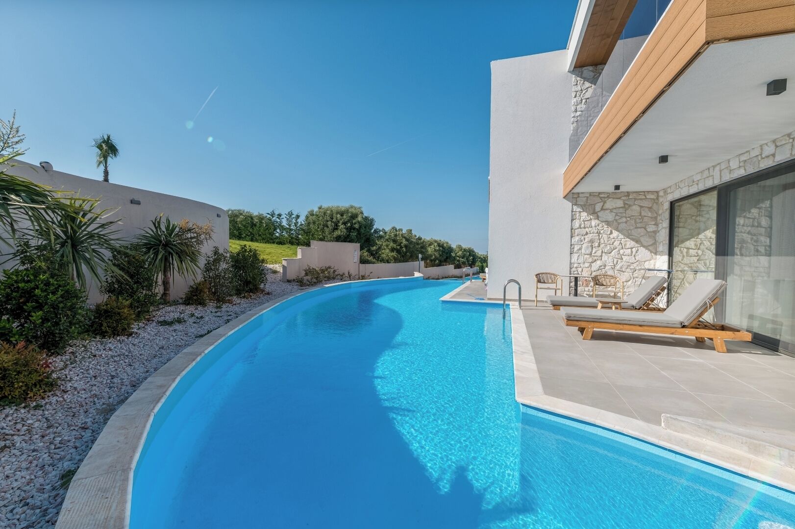 A modern swimming pool with clear blue water, surrounded by a white wall and a stone facade.