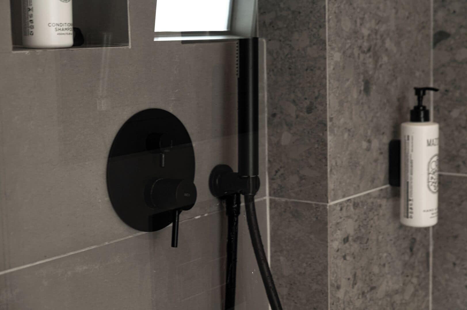 A bathroom with a modern shower control panel, a handheld showerhead, and a bottle of shampoo on the wall.