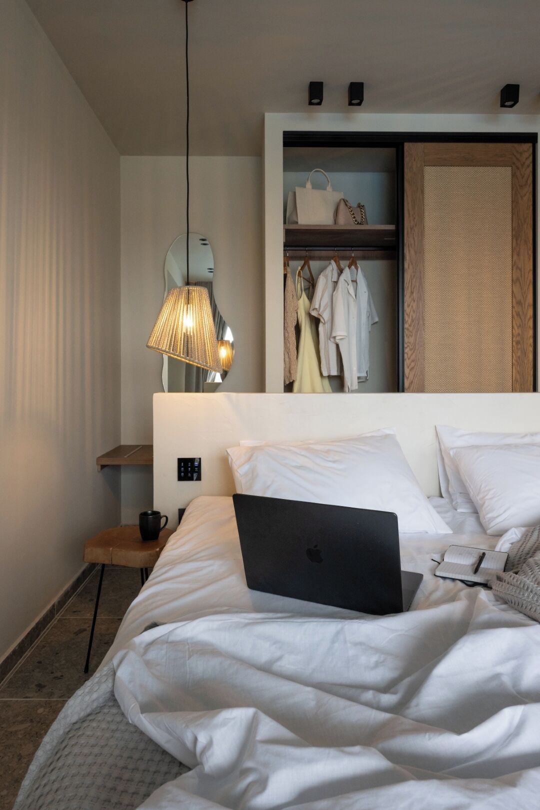 A neatly made bed with a laptop, a bedside table with a mug, and a hanging lamp.