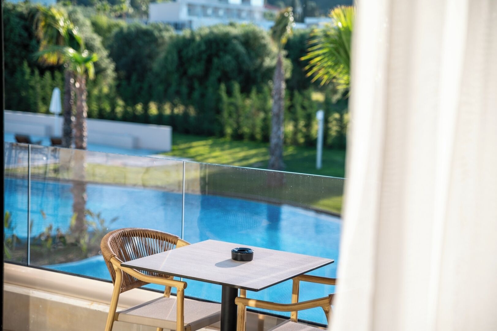 A balcony with a glass railing overlooking a swimming pool, a wicker chair, and a small table with a cup.