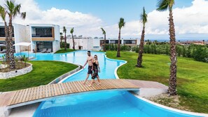 A swimming pool with a wooden deck, surrounded by palm trees and a modern building with large windows. - Anna Island (Pefkochori)