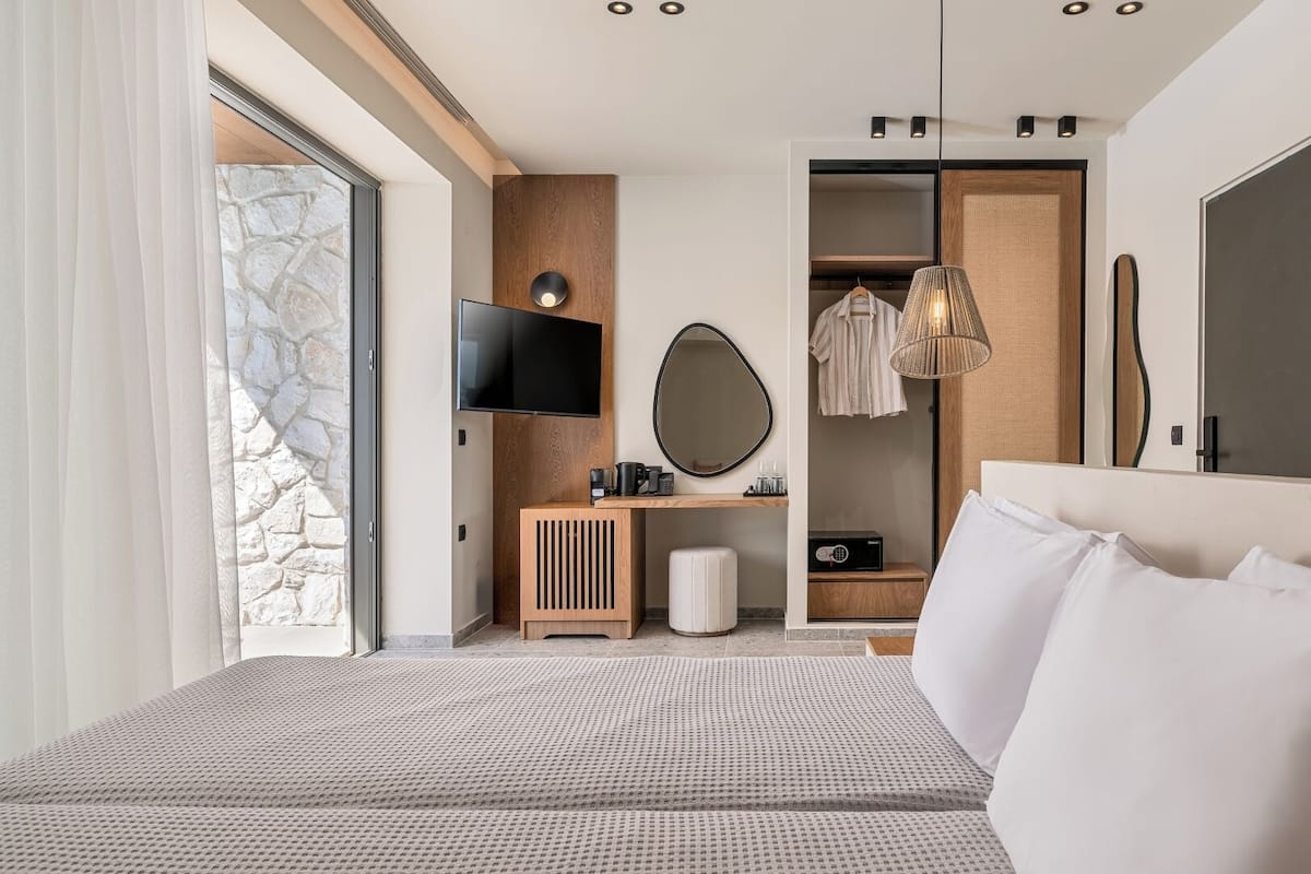 A modern hotel room with a large bed, a TV, a mirror, a wardrobe, and a desk with a coffee maker.
