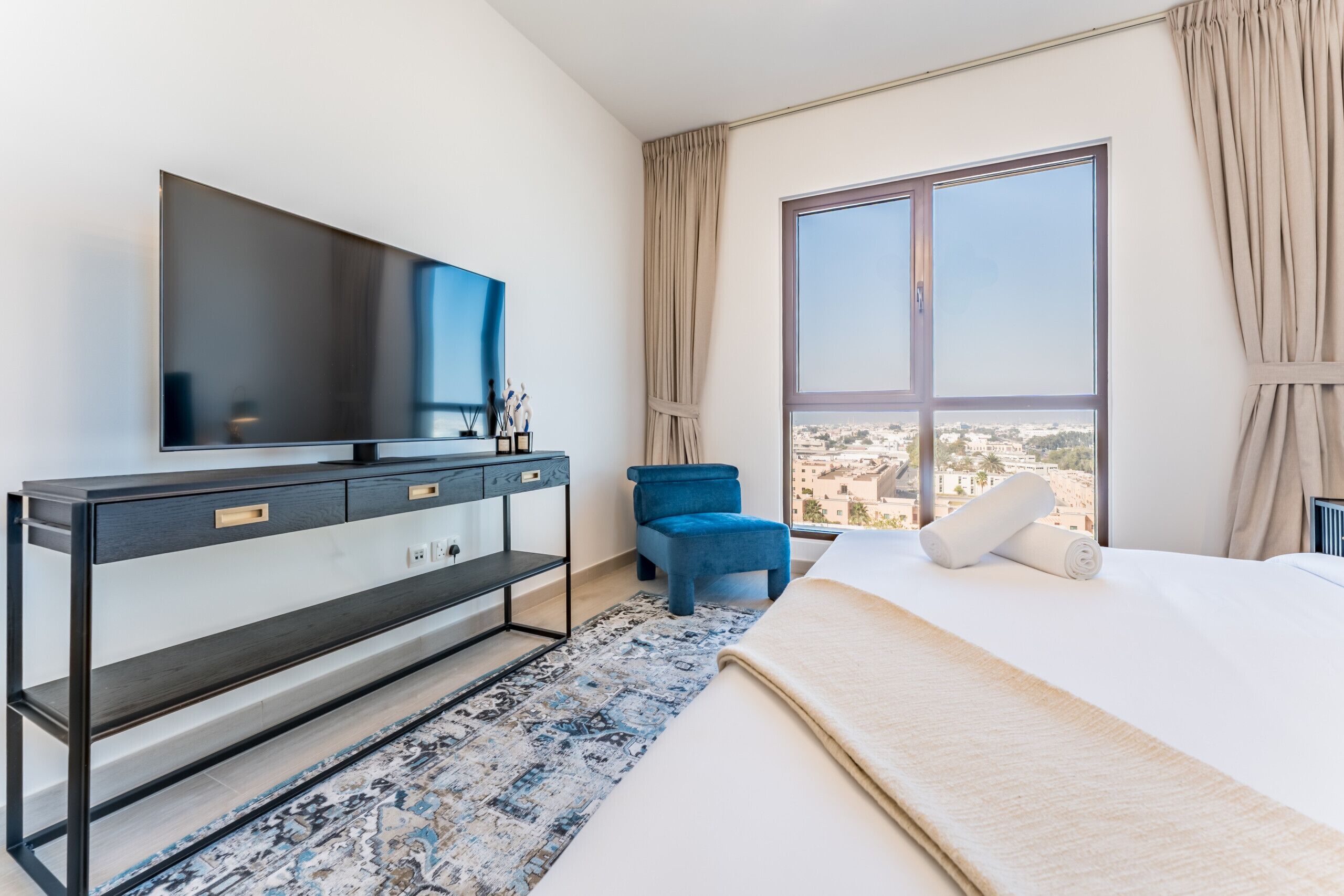 Ein modernes Hotelzimmer mit einem großen Bett, einem flachbildfernseher an der Wand, einem blauen Sessel und Blick auf die Stadt durch ein großes Fenster.