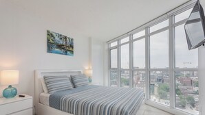 A modern bedroom with a large window, a bed with striped bedding, a nightstand with a lamp, and a painting on the wall. - 1BR Apartment in Toronto close to CN tower and Rogers Centre (Toronto)