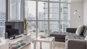A modern living room with a large TV, a grey sofa, and a view of a cityscape through floor-to-ceiling windows. - 1BR Apartment in Toronto close to CN tower and Rogers Centre (Toronto)