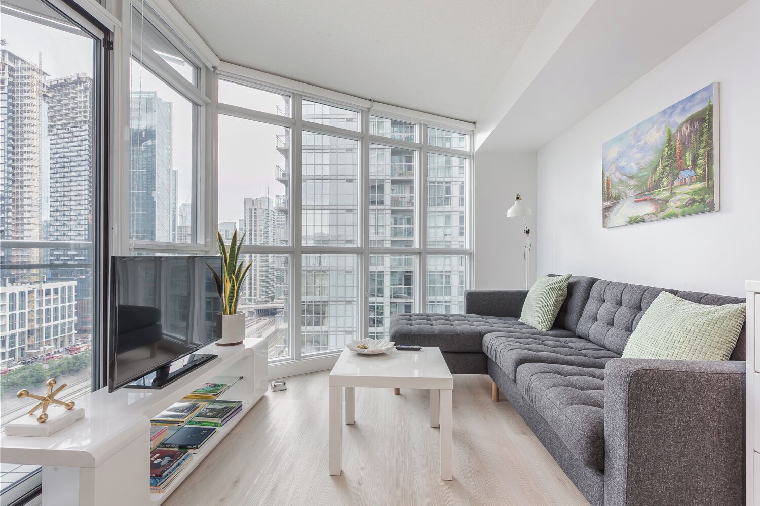 A modern living room with a grey sofa, a white coffee table, and a large window offering a city view.