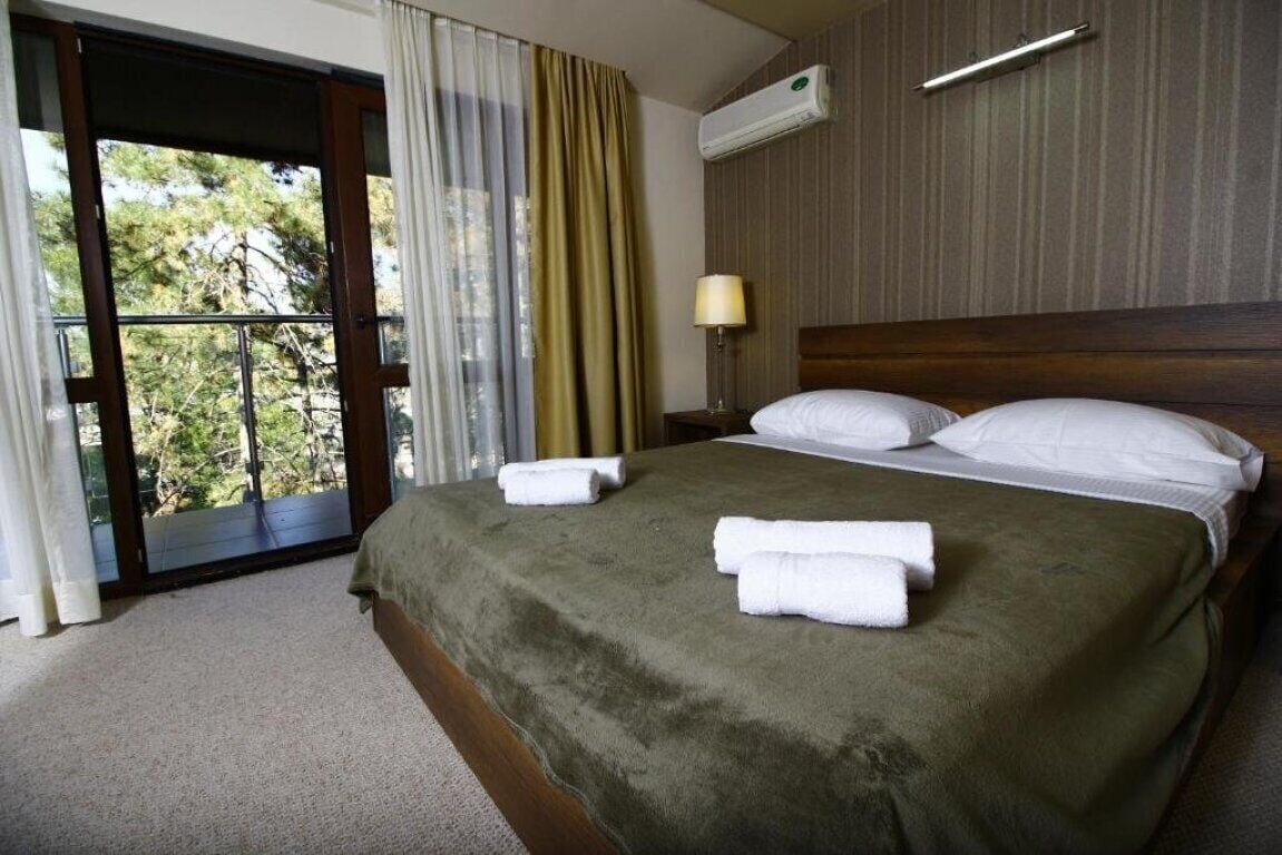 A hotel room with a large bed, a window with curtains, an air conditioning unit, and a nightstand with a lamp.