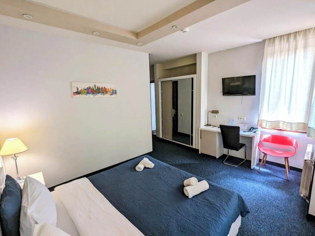 A hotel room with a bed, a desk, a chair, a TV, and a window with curtains.