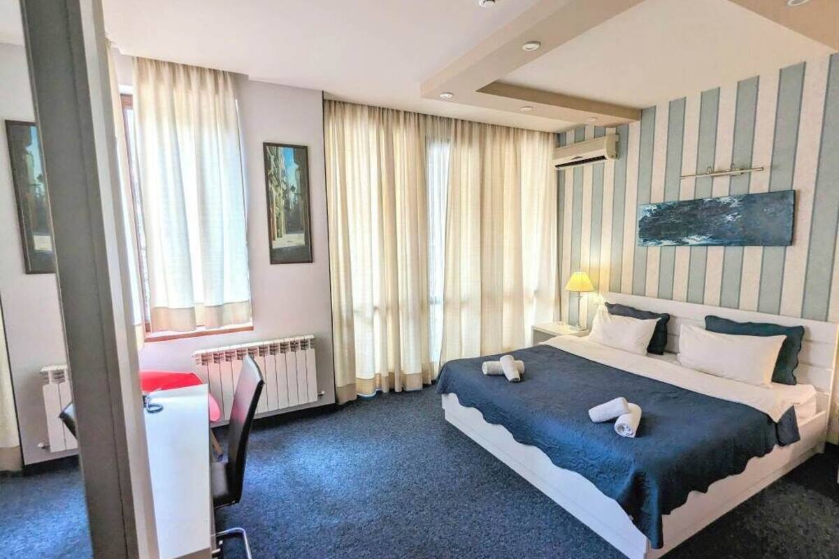 A hotel room with a large bed, a desk with a chair, a radiator, and a window with curtains.