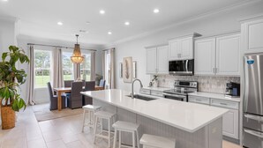 A modern kitchen with white cabinetry, a large island with stools, stainless steel appliances, and a dining area with a view of the outdoors.
