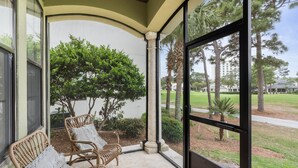 A covered porch with wicker chairs, a view of a landscaped area with trees and a building in the background.