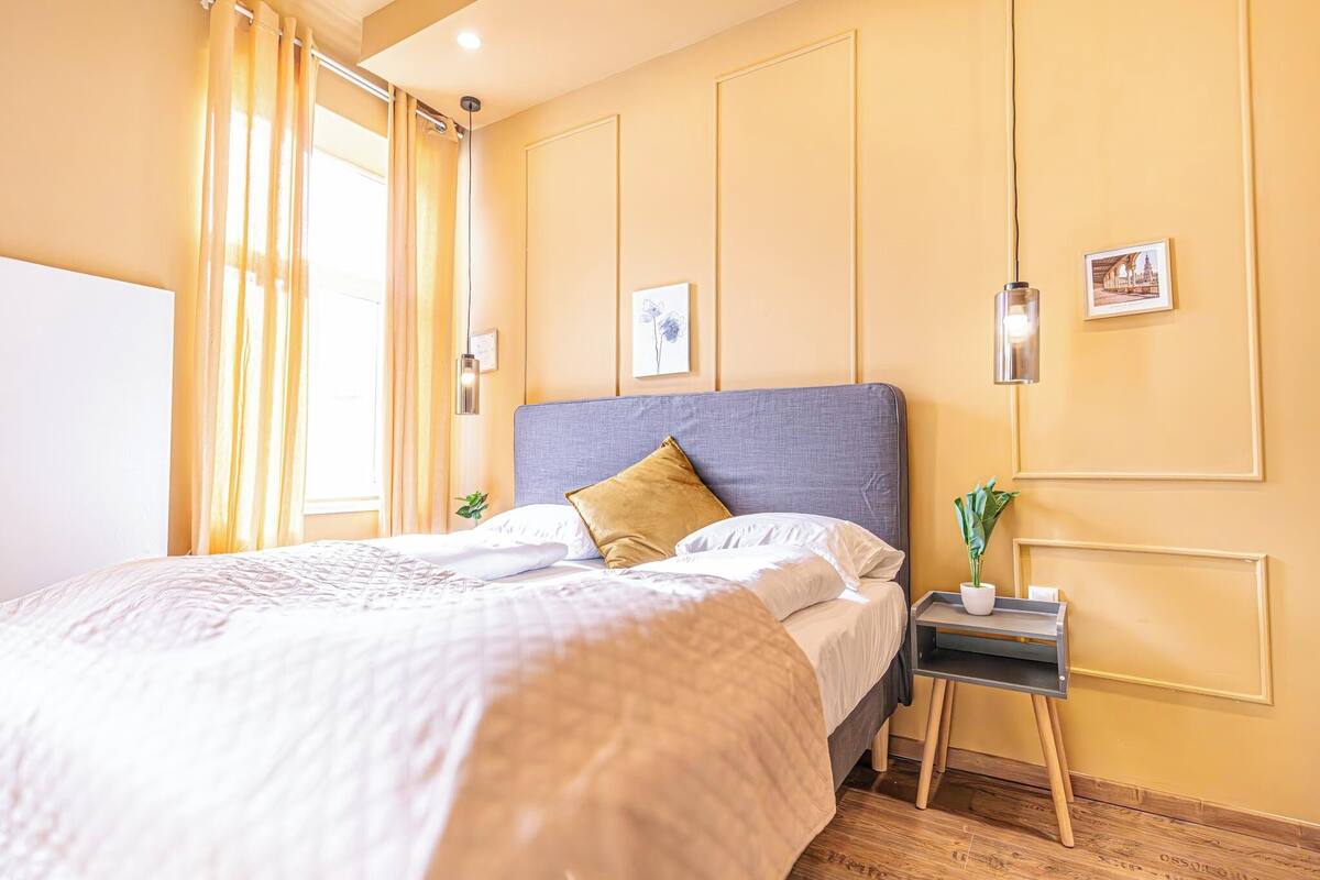 A neatly arranged bedroom with a large bed, a bedside table with a plant, and a wall-mounted light fixture.