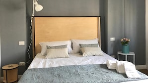 Egy gondosan megterített ágy, fafejtámlával, fehér ágyneművel és csíkos párnákkal. Mellette egy éjjeli lámpa, egy kis, kerek asztal, és egy virágos váz áll egy kisebb, mellékes asztalon.
