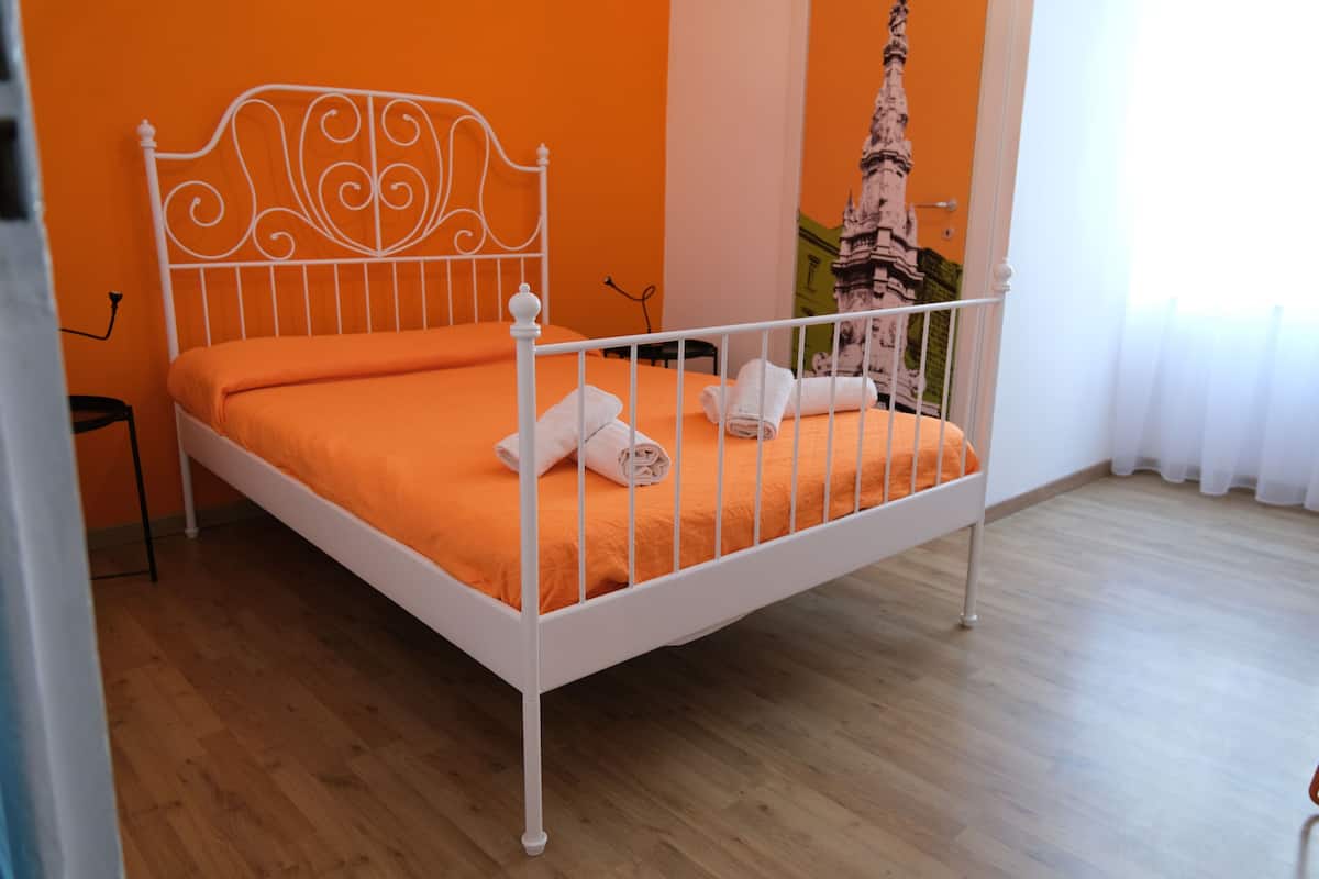 A neatly made bed with white metal headboard and orange bedding, a wooden floor, and a wall with a decorative picture.