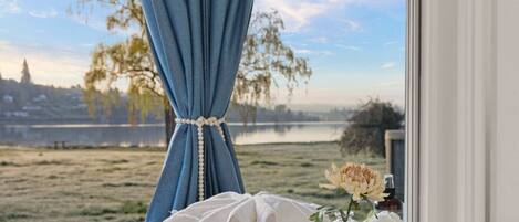 A room with a view of a lake, a window with blue curtains tied with a beaded string, a white pillow, a glass vase with flowers, and a wicker tray with a bottle.