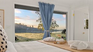 Une chambre avec une grande fenêtre, un lit, une table de chevet avec un panier, et une vue sur un lac et des arbres.