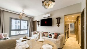 A modern living room with a round marble table set for dining, a large window, a flat-screen TV, and a chandelier. - Xuru Stays I Authentic Traditions Cozy Stays, 1bdr (Cairo)