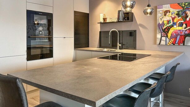 Een moderne keuken met een centraal eiland, barkrukken, een inbouwoven en een hanglamp.