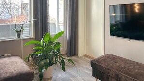 Een woonkamer met een groot raam, een kamerplant, een bank, een televisie en een houten vloer.