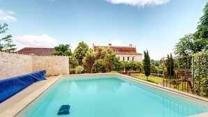 A rectangular swimming pool with a clear blue water surface, surrounded by a stone wall and a metal fence. In the background, there is a large building with a red-tiled roof and a well-maintained garden.