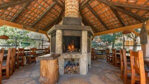 An outdoor patio with a stone fireplace, wooden chairs, and a thatched roof structure.