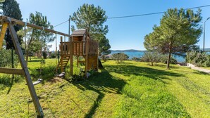 A wooden play structure in a grassy yard with a clear view of the sea and mountains in the background.