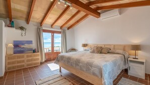 Ein Schlafzimmer mit Holzbalkendecke, einem Bett mit gemusterten Textilien, einem Nachttisch und Meerblick durch eine Tür.