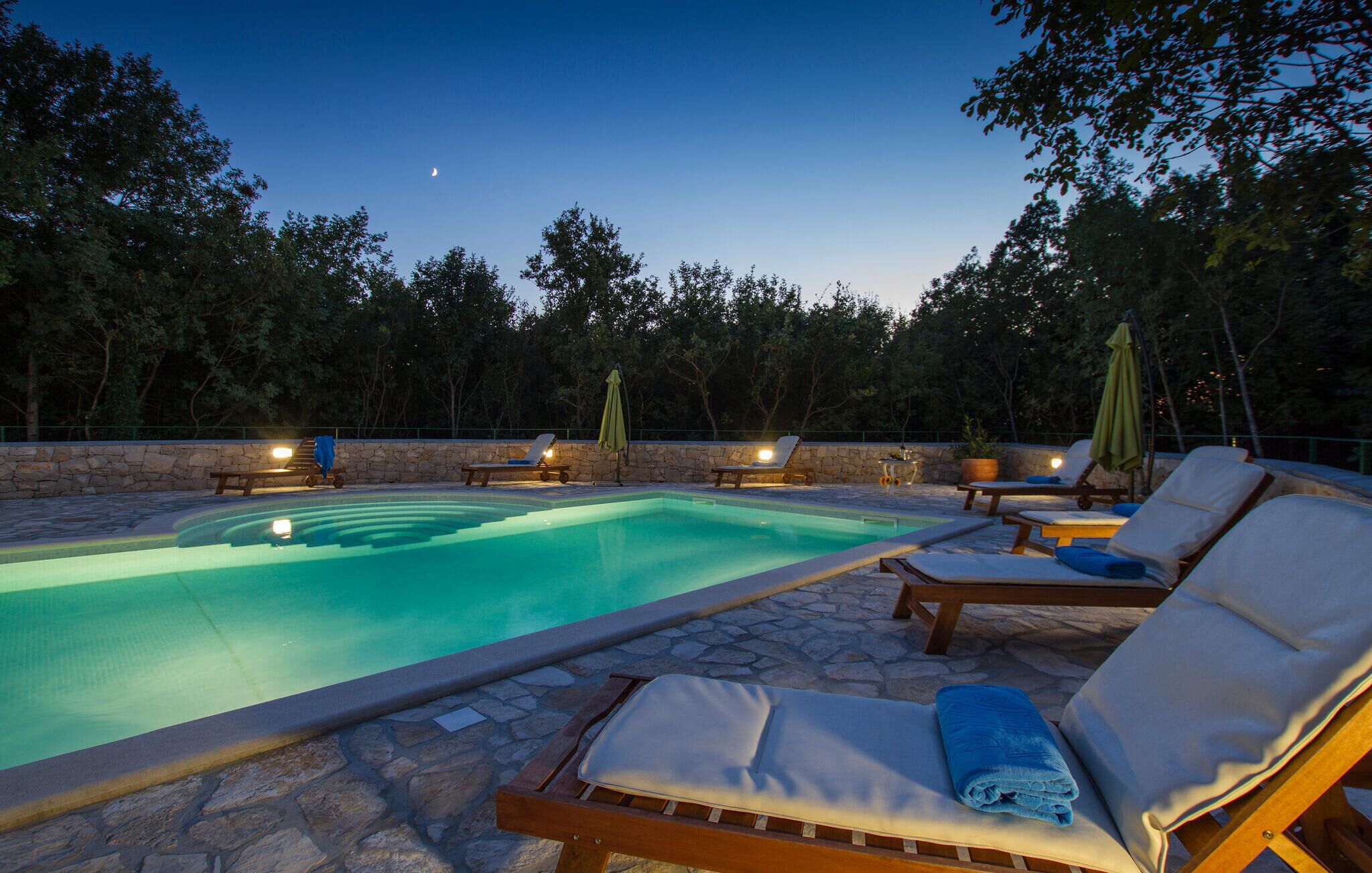 A swimming pool with lounge chairs and umbrellas, surrounded by stone walls and trees.