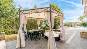 A covered outdoor seating area with a table and chairs, surrounded by greenery and a pool in the background. - Cozy home in Capolona (Capolona)
