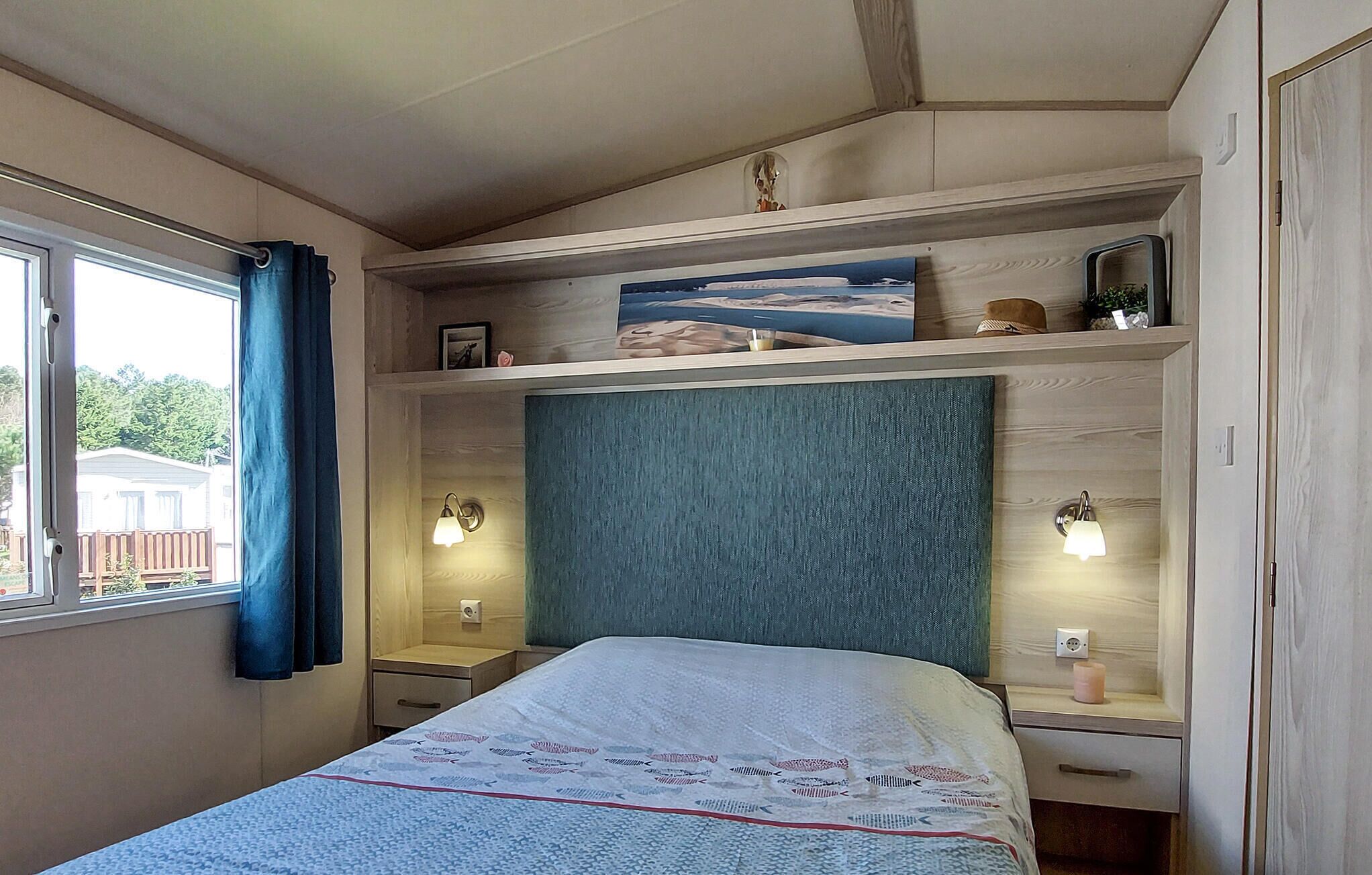 A bedroom with a bed, a nightstand, a shelf with decorative items, and a window with curtains.