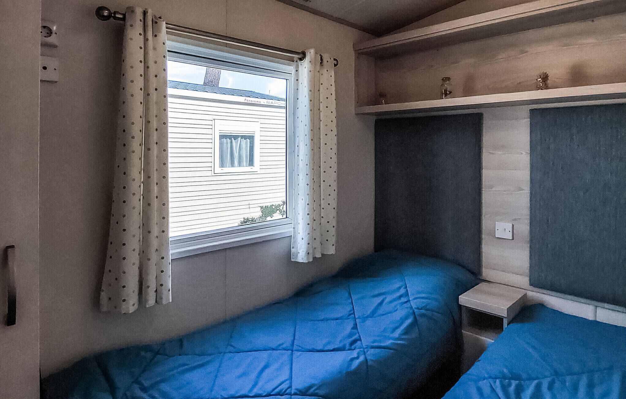A small, compact room with two single beds, a window with curtains, a shelf with a small item, and a wall socket.