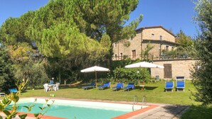 Zona de piscina con sillas de descanso y sombrillas, rodeada de vegetación y un edificio de piedra.