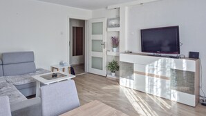 Una sala de estar moderna con un sofá gris, una mesita con un control remoto, un televisor sobre un mueble de madera y una puerta que da a otra habitación.