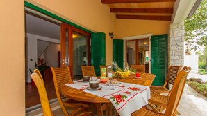 A wooden table with a white tablecloth, surrounded by wooden chairs, set on a patio with green shutters and a stone wall. - Gorgeous home in Koromacno with WiFi (Koromacno)