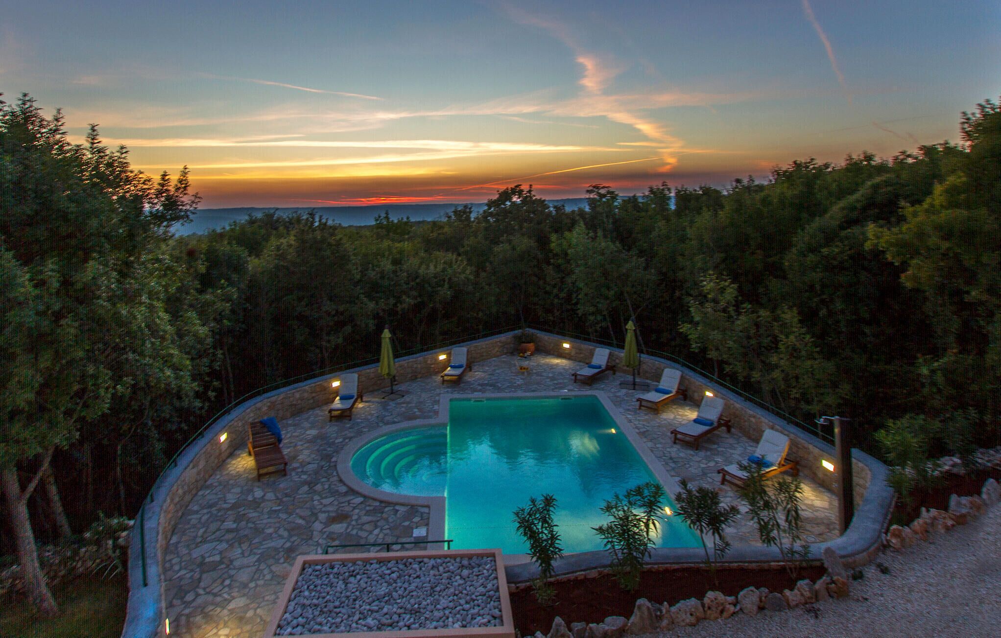 A pool area with lounge chairs and a stone deck, surrounded by trees and overlooking a sunset view.