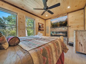 A wooden cabin bedroom with a large bed, a ceiling fan, a fireplace, and a scenic view through the windows.