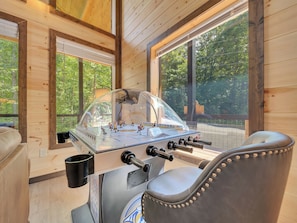 A wooden-floored room with a foosball table, a window with a view of trees, and a chair.