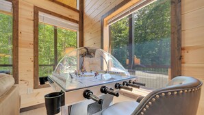 Une pièce au plancher en bois, avec un baby-foot, une fenêtre donnant sur des arbres et une chaise.