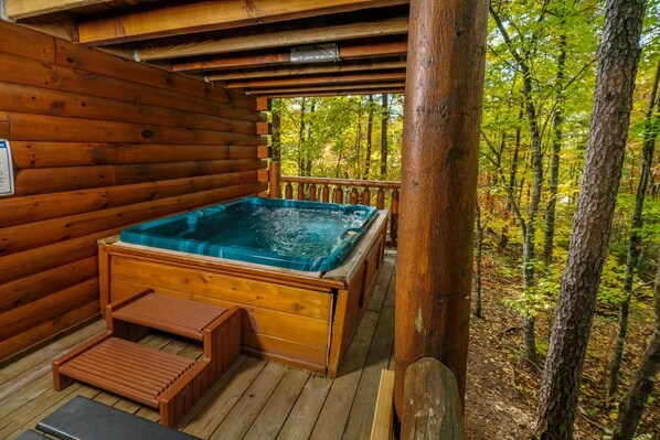 A wooden cabin with a hot tub on a deck surrounded by trees.