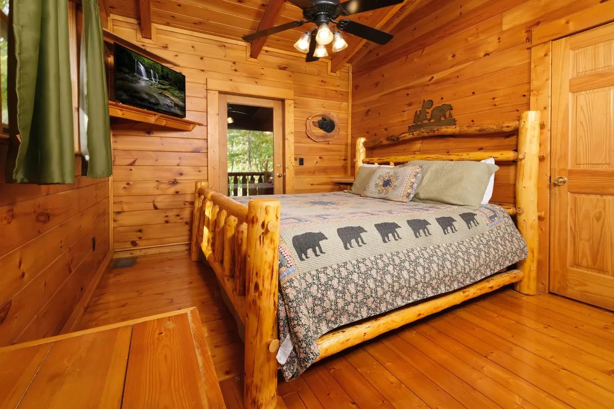 Ein Zimmer in einer Holzhütte mit einem Bett, einem Deckenventilator, einem Wandfernseher und einer Tür, die nach draußen führt.
