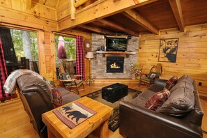 A cozy cabin interior with a stone fireplace, leather furniture, and a large window overlooking the outdoors.