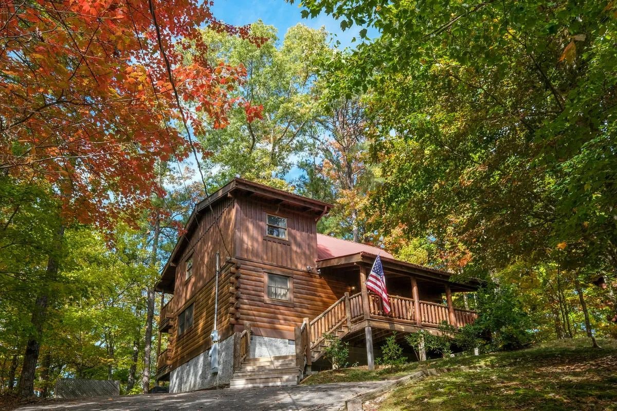 A wooden cabin with a porch and a staircase, surrounded by autumn foliage.