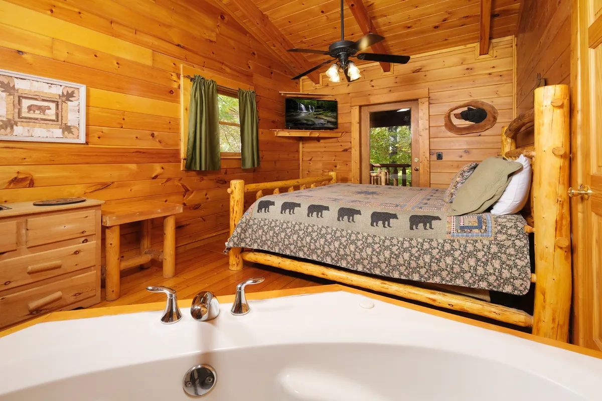 A wooden cabin bedroom with a bed, a desk, a television, and a bathroom with a bathtub.