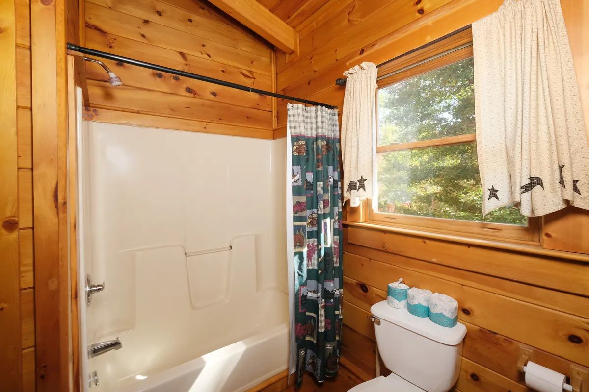 A wooden cabin bathroom with a shower, toilet, and window with curtains.